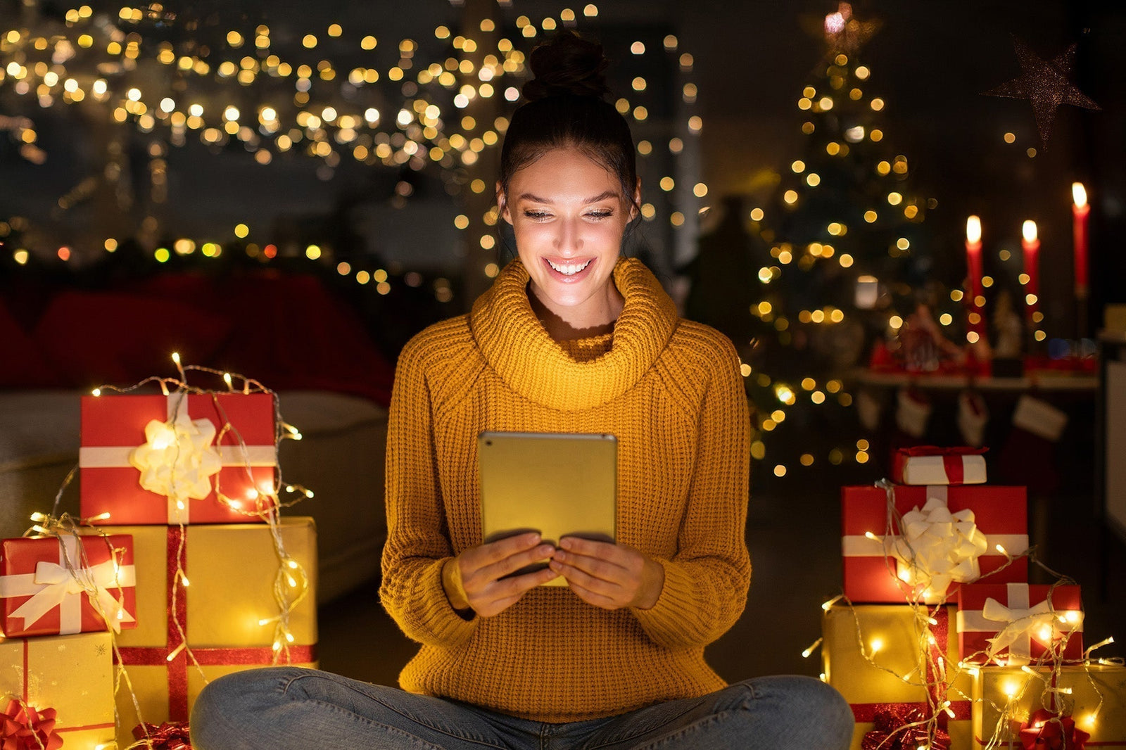 Woman looking at a tablet sitting in front of holiday lights and gifts
