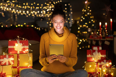 Woman looking at a tablet sitting in front of holiday lights and gifts
