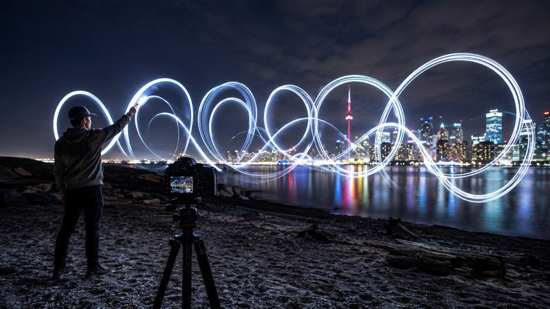 Photographer creating looping light graffiti beside a tripod-mounted camera and city skyline at night.