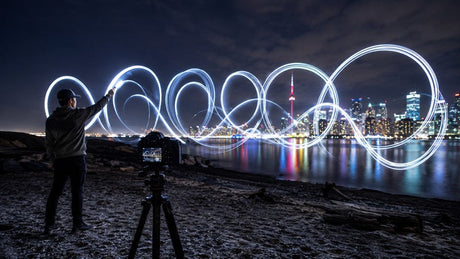 Photographer creating looping light graffiti beside a tripod-mounted camera and city skyline at night.
