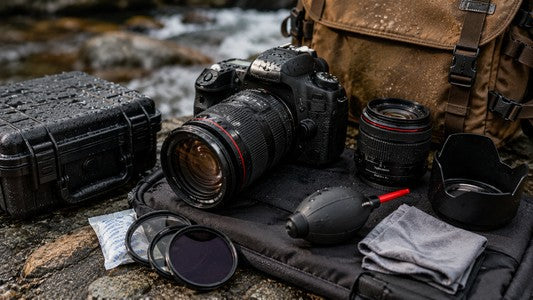 Weather-resistant DSLR camera gear arranged with a protective case and cleaning accessories beside a rocky stream.
