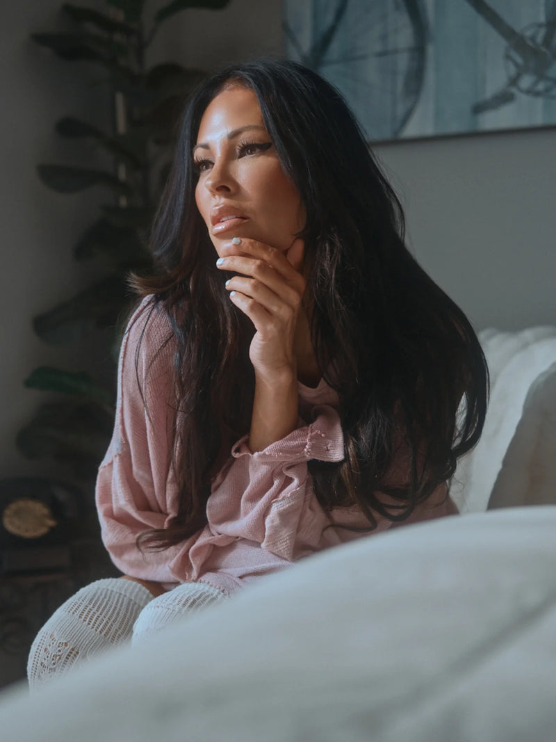 Woman in a pink shirt sitting on a bed in a softly lit room.