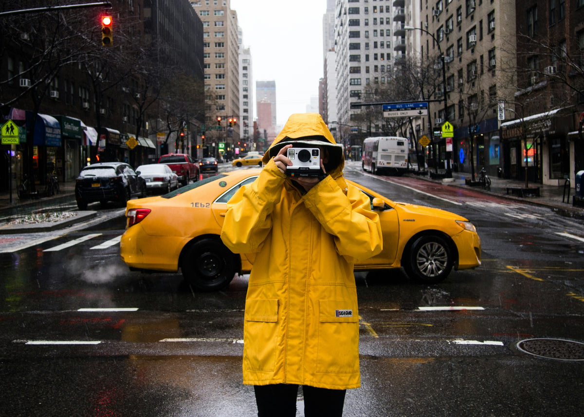 Person in a yellow raincoat taking a photo on a city street with a yellow taxi in the background.