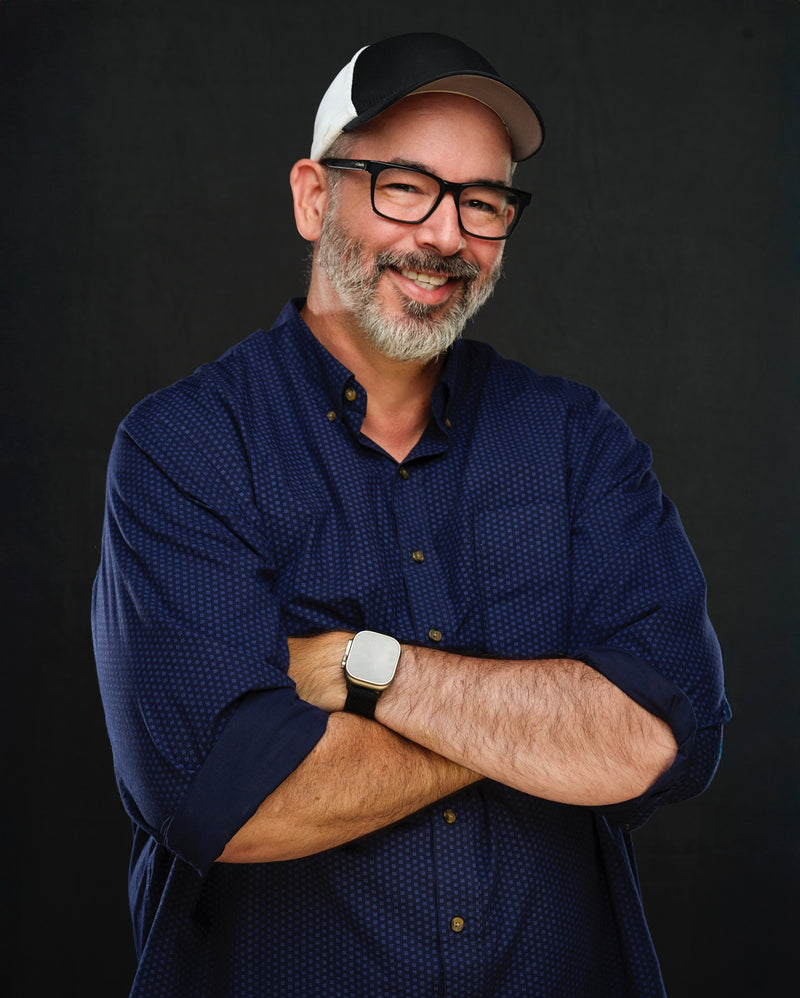 Man wearing a blue shirt, black cap, and glasses with arms crossed against a dark background