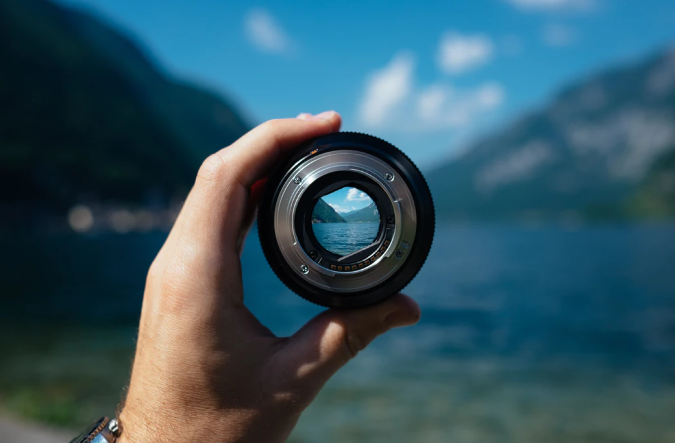 Hand holding a camera lens. In the background is water surrounded by mountanis. 